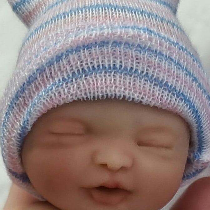 May include: A small, nude baby doll wearing a pink and blue striped hat and a white diaper. The doll is sleeping with its eyes closed.