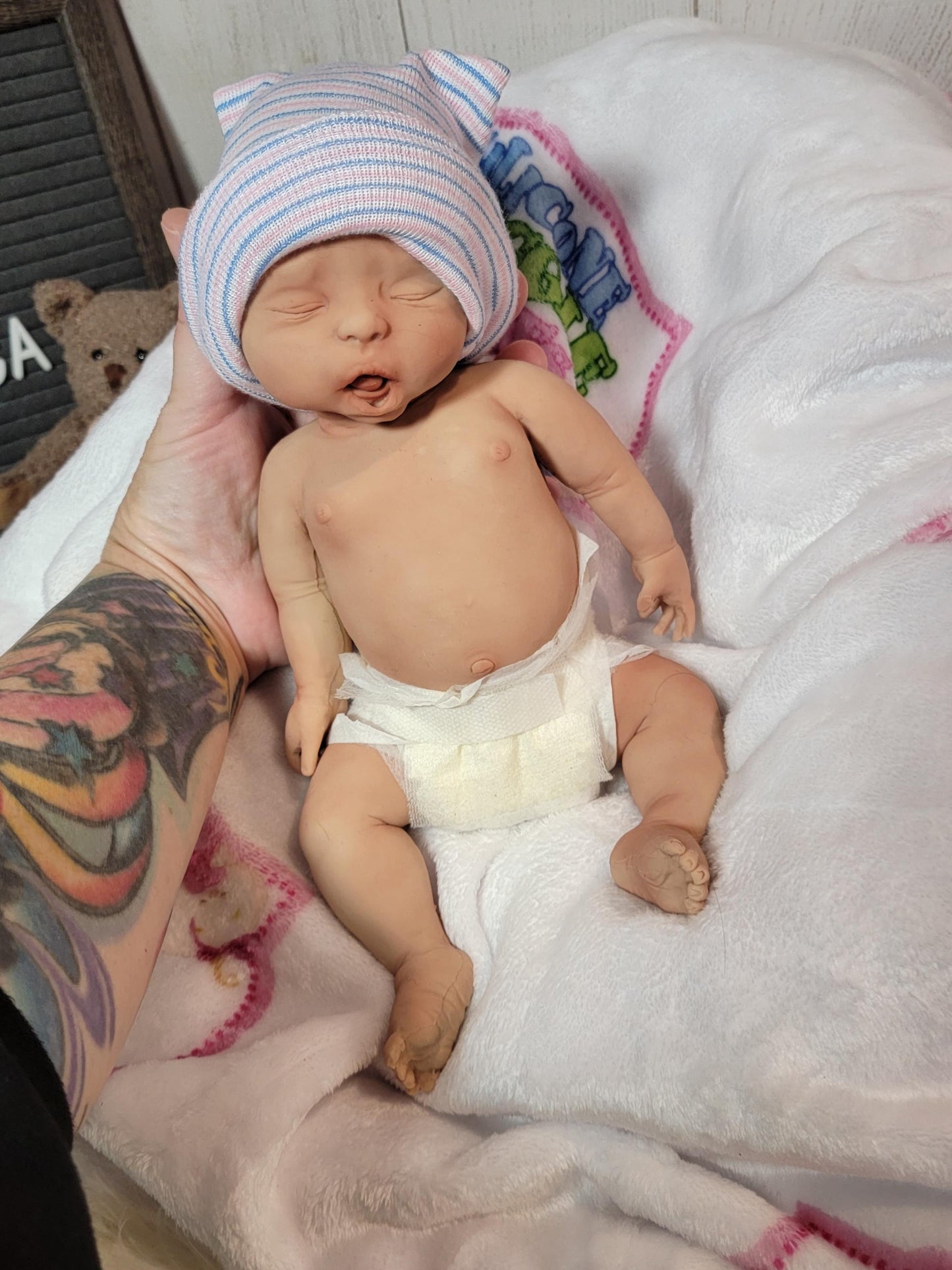 May include: A realistic newborn baby doll with light skin, closed eyes, and an open mouth. The doll wears a white diaper and a soft knit hospital cap featuring horizontal blue and pink stripes. The doll has detailed facial features and limbs.