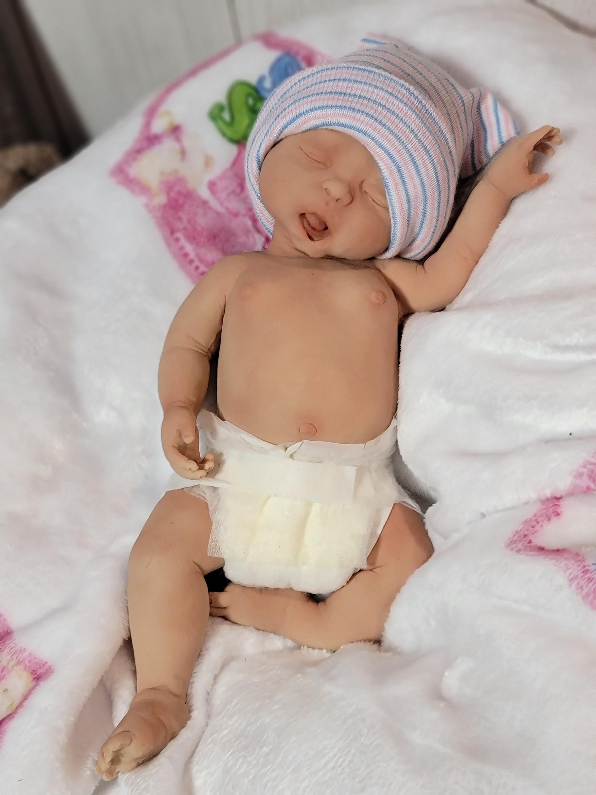 May include: A realistic newborn baby doll with light skin, closed eyes, and an open mouth. The doll wears a white diaper and a soft, knitted hospital cap featuring thin horizontal stripes in light blue and pink, resting on a white blanket.